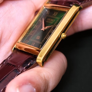 May include: A gold-toned Seiko Quartz watch with a rectangular green face and Roman numeral hour markers. The watch has a brown leather band. The watch face displays the Seiko Quartz logo. The watch is a classic timepiece.