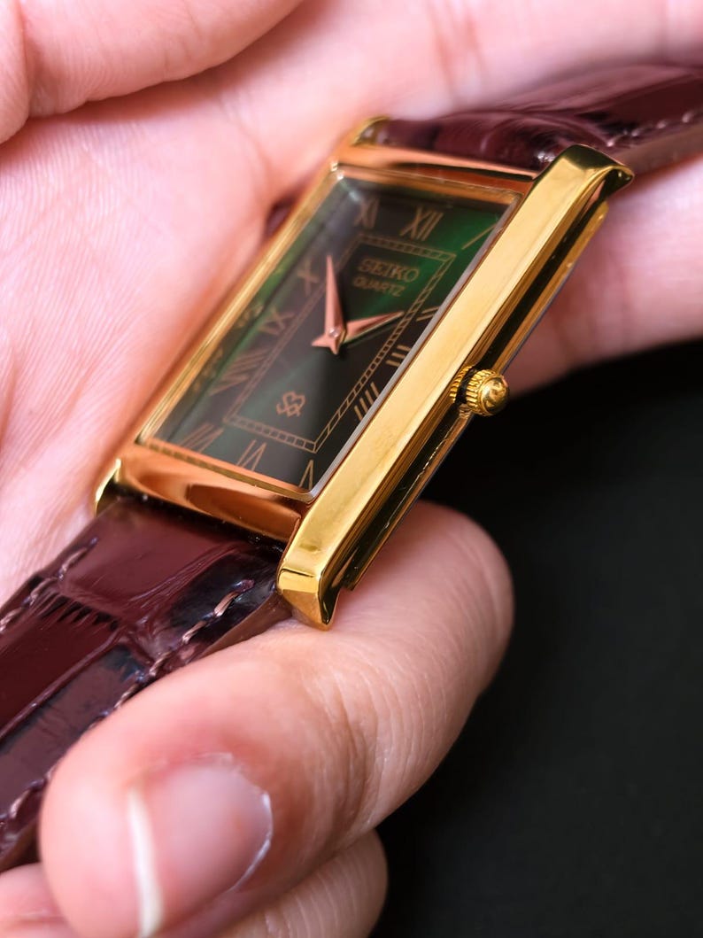 May include: A gold-toned Seiko Quartz watch with a rectangular green face and Roman numeral hour markers. The watch has a brown leather band. The watch face displays the Seiko Quartz logo. The watch is a classic timepiece.