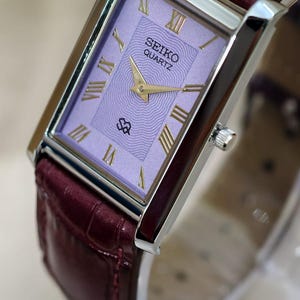 Peut inclure: Montre Seiko Quartz rectangulaire avec un cadran lavande et des chiffres romains dorés. Le bracelet est en cuir bordeaux. Le cadran de la montre porte le logo Seiko Quartz et un motif texturé. Le boîtier de la montre est argenté.