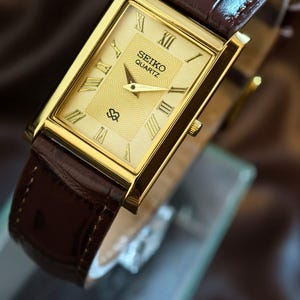 Puede incluir: Reloj Seiko Quartz dorado con esfera rectangular y números romanos. El reloj tiene una esfera texturizada y una correa de cuero marrón. El reloj se muestra sobre una superficie transparente sobre un fondo borroso.