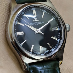 May include: A stainless steel Jaeger-LeCoultre Club watch with a black dial and silver hands. The watch has a green leather strap. The dial features the Jaeger-LeCoultre logo and the text "MADE T SWISS".