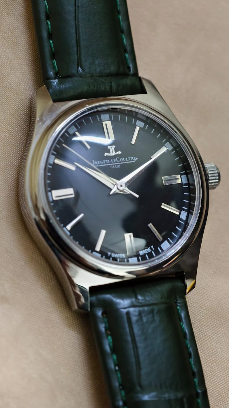 May include: A stainless steel Jaeger-LeCoultre Club watch with a black dial and silver hands. The watch has a green leather strap. The dial features the Jaeger-LeCoultre logo and the text "MADE T SWISS".