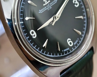 Può includere: Primo piano di un orologio da polso argentato con quadrante verde scuro e cinturino in pelle verde. Il quadrante dell'orologio presenta lancette e indicatori argentati, con il logo Jaeger-LeCoultre Club. L'orologio è angolato.