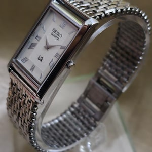 May include: A silver Seiko Quartz watch with a rectangular face and a woven metal band. The watch face is white with black Roman numerals and hands. The band is made of small, interconnected metal links.