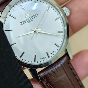 May include: A silver Jaeger-LeCoultre Club watch with a white face and brown leather strap. The watch has silver hands and hour markers. The words "JAEGER-LECOULTRE CLUB" are printed on the face. The watch is marked "SWISS MADE".