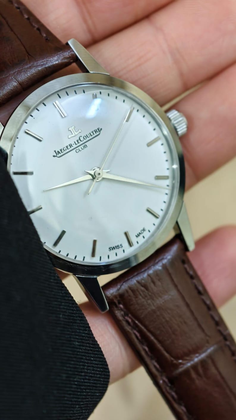 May include: A silver Jaeger-LeCoultre Club watch with a white face and brown leather strap. The watch has silver hands and hour markers. The words "JAEGER-LECOULTRE CLUB" are printed on the face. The watch is marked "SWISS MADE".