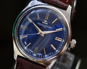 Può includere: Un orologio Jaeger-LeCoultre Club argentato con quadrante blu scuro e lancette e indici dorati. L'orologio ha un cinturino in pelle marrone. Il quadrante dell'orologio riporta la scritta "JAEGER-LECOULTRE CLUB" e "T SWISS MADE T".