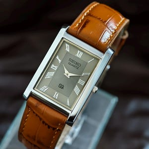 Könnte beinhalten: Eine rechteckige Seiko Quartz-Uhr mit silbernem Gehäuse und braunem Lederarmband. Das Zifferblatt der Uhr ist grau mit silbernen römischen Ziffern und Zeigern. Das Armband hat ein Krokodilmuster.