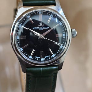 May include: A silver-toned Jaeger-LeCoultre Club watch with a black face and silver hands. The watch has a dark green leather band. The watch face displays the Jaeger-LeCoultre logo and the words "CLUB", "SWISS", and "MADE".