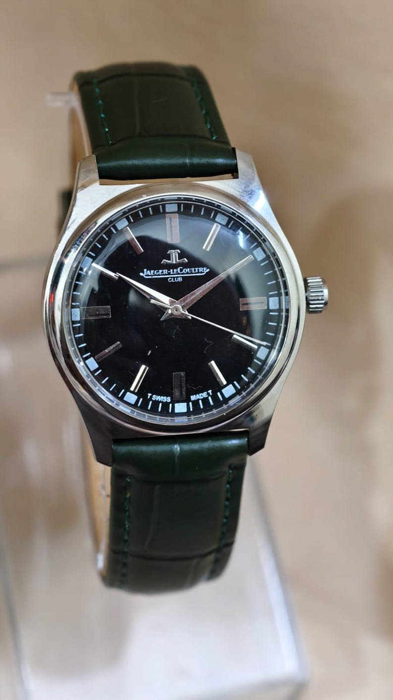 May include: A silver-toned Jaeger-LeCoultre Club watch with a black face and silver hands. The watch has a dark green leather band. The watch face displays the Jaeger-LeCoultre logo and the words "CLUB", "SWISS", and "MADE".