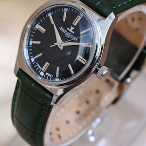 May include: A silver-toned wristwatch with a black face and green leather band. The watch face features silver hands and markers, with the brand name "Jaeger-LeCoultre" visible. The green band is textured.