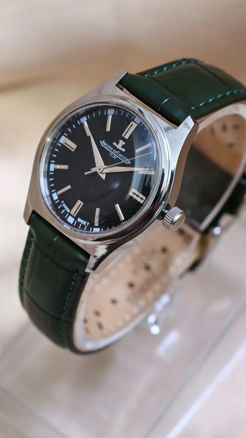 May include: A silver-toned wristwatch with a black face and green leather band. The watch face features silver hands and markers, with the brand name "Jaeger-LeCoultre" visible. The green band is textured.