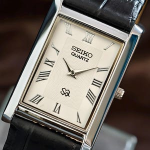 Puede incluir: Reloj Seiko Quartz rectangular con caja plateada y esfera color crema. La esfera presenta números romanos negros, agujas negras y el logotipo de Seiko Quartz. La correa es de cuero negro.