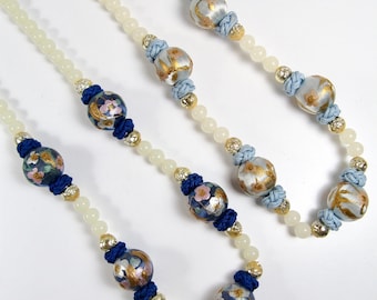 Puede incluir: Un collar largo con cuentas blancas y cuentas con patrones azules y dorados. Las cuentas tienen un diseño floral.
