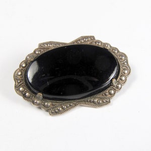 May include: A black oval cabochon gemstone set in a silver tone ornate frame. The frame is decorated with small beads and a scalloped edge.