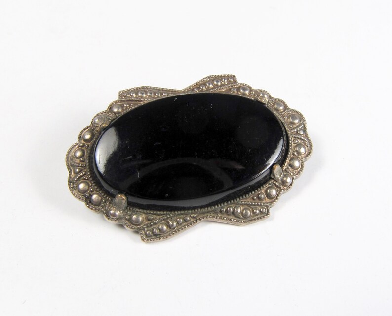 May include: A black oval cabochon gemstone set in a silver tone ornate frame. The frame is decorated with small beads and a scalloped edge.