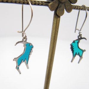 May include: A pair of silver earrings with a turquoise blue goat design. The earrings are hanging from a display stand.