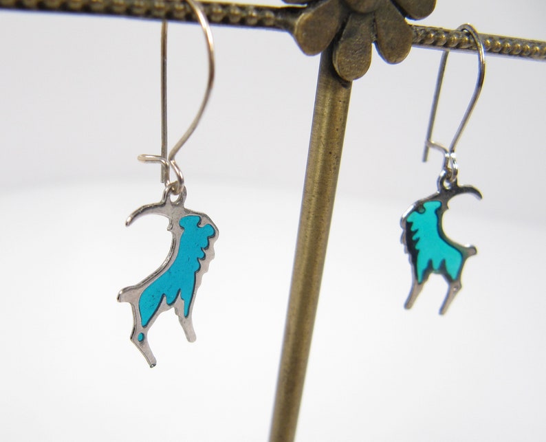 May include: A pair of silver earrings with a turquoise blue goat design. The earrings are hanging from a display stand.