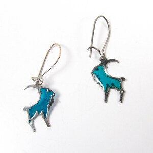 May include: A pair of silver earrings with a blue enamel goat design. The goat is outlined in silver and has a blue enamel fill.
