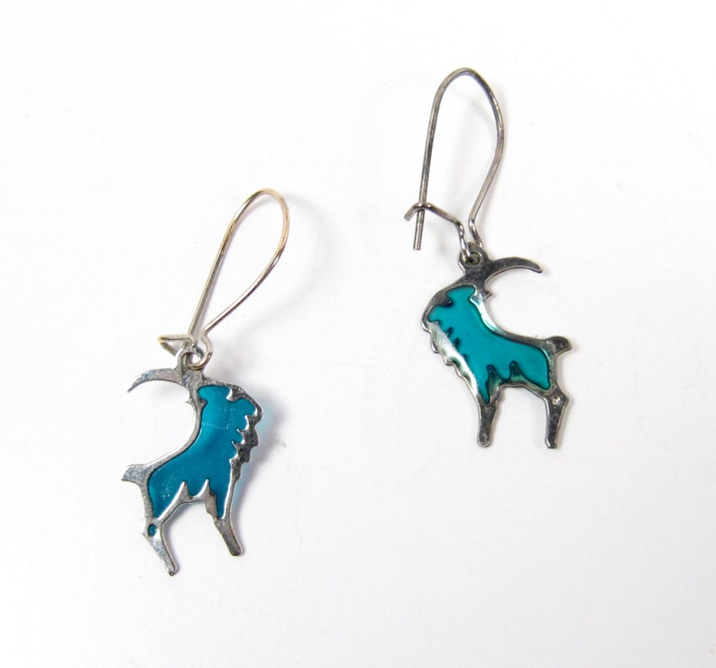 May include: A pair of silver earrings with a blue enamel goat design. The goat is outlined in silver and has a blue enamel fill.
