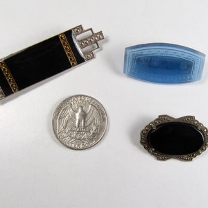 May include: Three vintage brooches featuring black and blue stones. The brooches are silver toned and have intricate designs. The brooches are approximately the size of a 25 pence coin.
