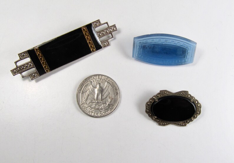 May include: Three vintage brooches featuring black and blue stones. The brooches are silver toned and have intricate designs. The brooches are approximately the size of a 25 pence coin.