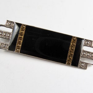 May include: A black enamel brooch with gold accents and a geometric design. The brooch is made of silver and has a rectangular shape.