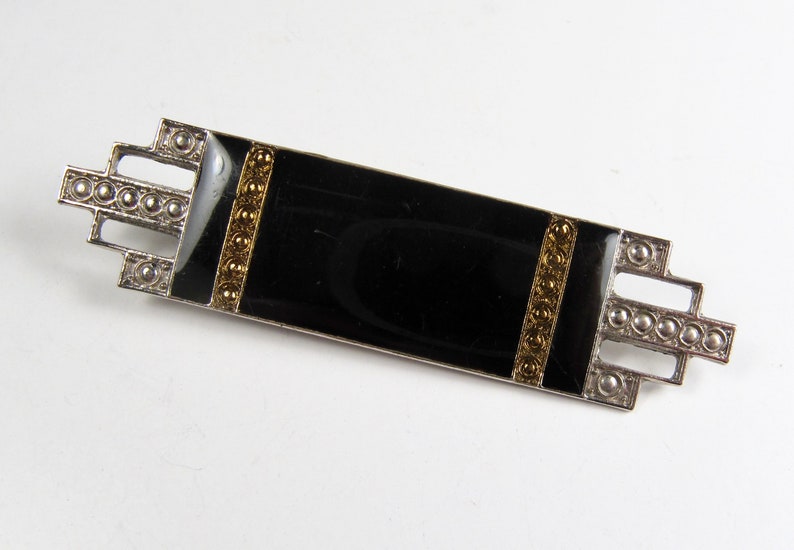May include: A black enamel brooch with gold accents and a geometric design. The brooch is made of silver and has a rectangular shape.