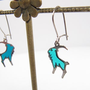 May include: A pair of silver dangle earrings with a turquoise blue goat design.