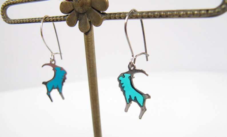 May include: A pair of silver dangle earrings with a turquoise blue goat design.