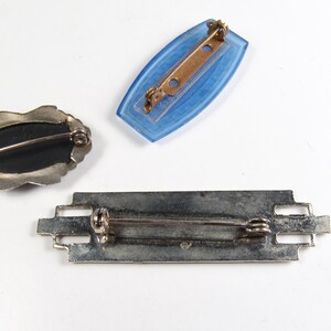 May include: Three vintage brooches. One is a silver-toned oval with a black centre. Another is a blue glass rectangle. The third is a silver-toned rectangular bar with an intricate design.