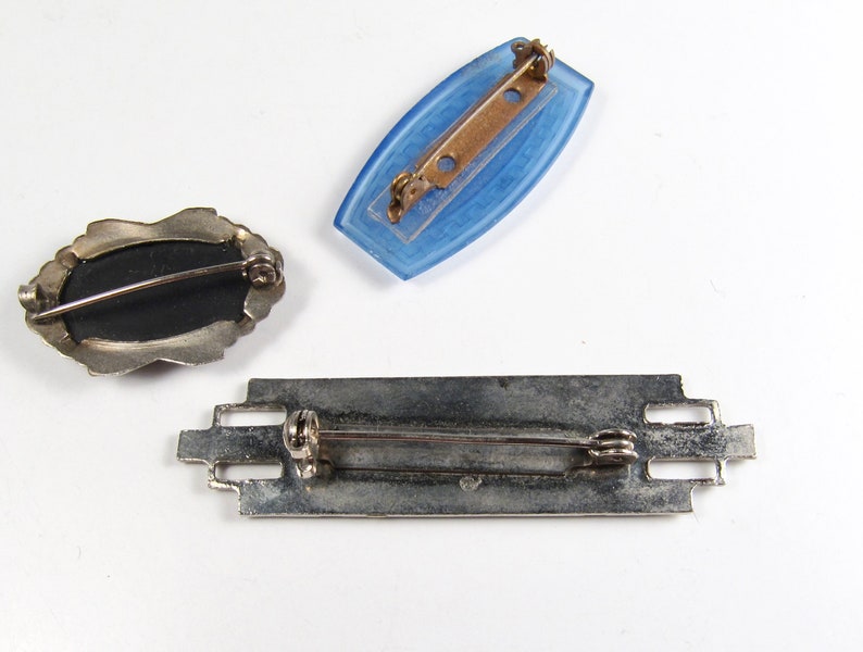 May include: Three vintage brooches. One is a silver-toned oval with a black centre. Another is a blue glass rectangle. The third is a silver-toned rectangular bar with an intricate design.