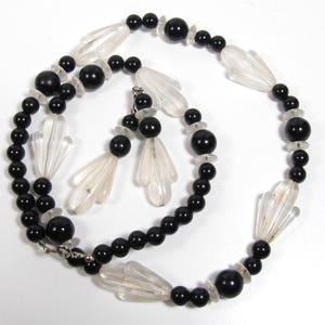 以下が含まれることがあります： 黒と透明なガラスビーズのネックレスとイヤリングのセット。ネックレスは黒い丸いビーズと扇形の透明なガラスビーズでできています。イヤリングは扇形の透明なガラスビーズのペアです。