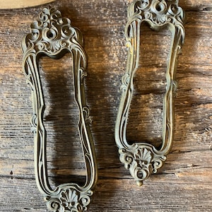 May include: Two ornate, antique-looking, brass door handles with intricate floral designs. The handles are in a vintage style and have a worn, aged finish.