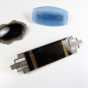 May include: Three vintage brooches with black enamel and silver tone metal. One brooch features a black oval stone, another has a rectangular shape with gold accents, and the third is a blue glass rectangle.
