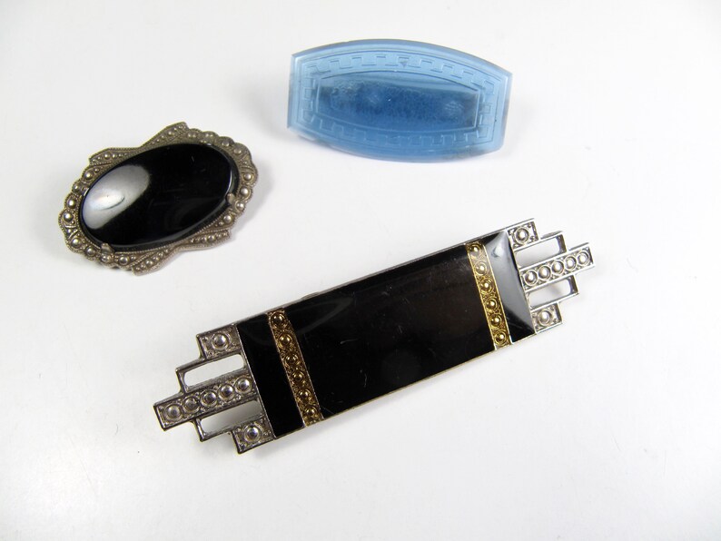 May include: Three vintage brooches with black enamel and silver tone metal. One brooch features a black oval stone, another has a rectangular shape with gold accents, and the third is a blue glass rectangle.