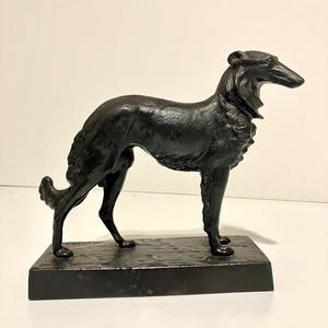 May include: A black sculpture of a Borzoi dog in a standing pose. The dog has a long, slender body and a long, narrow snout. The sculpture is mounted on a rectangular base.