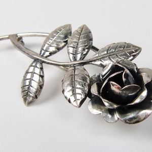 May include: A silver-coloured brooch depicting a rose and leaves. The brooch shows a detailed rose bloom with multiple petals and several textured leaves along a curved stem. The metal has a slightly aged appearance.