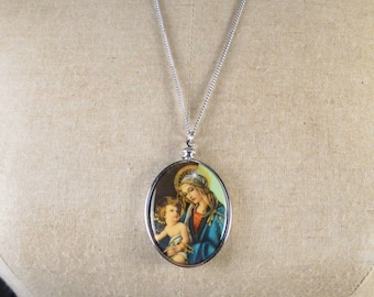 May include: A silver chain necklace with an oval pendant featuring a vintage image of the Virgin Mary holding a baby Jesus. The image is surrounded by a silver frame.
