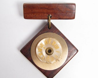 May include: A wooden brooch with a square shape and a round clock face in the centre. The clock face has Roman numerals and a gold-coloured rim. The brooch is attached to a rectangular wooden bar.