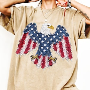 May include: Tan-colored t-shirt featuring a large graphic of an American bald eagle with outstretched wings. The eagle's wings are designed with red and white stripes, and the body is blue with white stars. The eagle has a white head and a yellow beak.