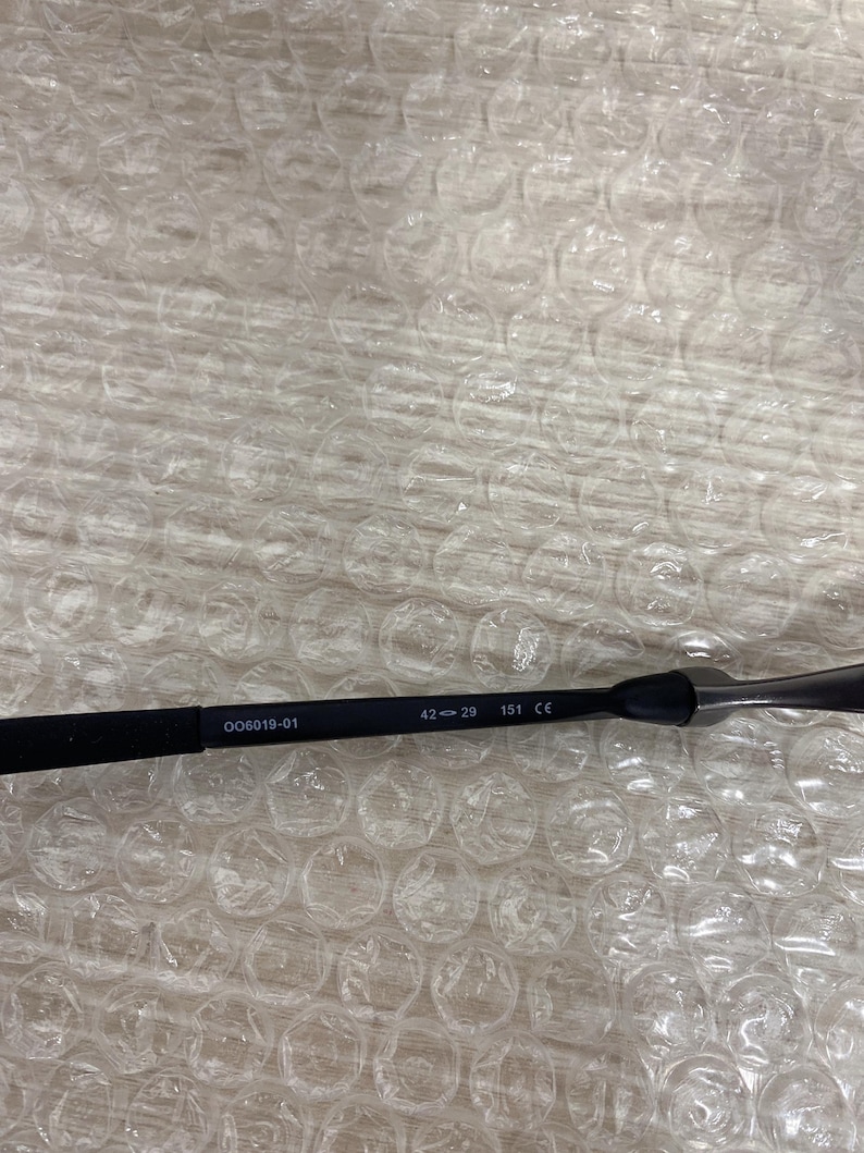 May include: Black eyeglasses frame with the text "006019-01", "42-29", and "151 CE". The frame is resting on a surface with a bubble wrap pattern.