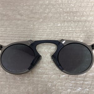 May include: Dark gray sunglasses with round lenses and a distinctive bridge design. The sunglasses have a sleek, modern aesthetic, with a dark frame and lenses. The brand name is visible on the left lens.