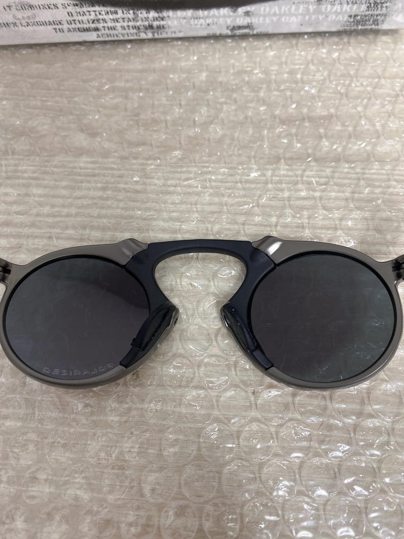 May include: Dark gray sunglasses with round lenses and a distinctive bridge design. The sunglasses have a sleek, modern aesthetic, with a dark frame and lenses. The brand name is visible on the left lens.