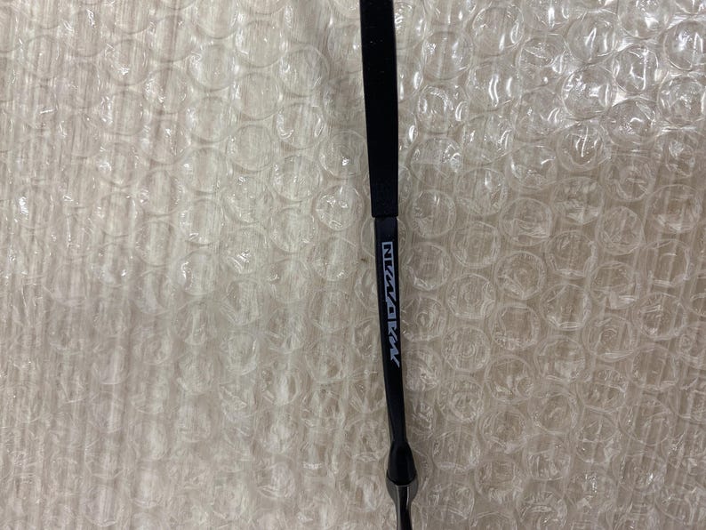 May include: A black eyeglass arm with the white text "NW/AKM" printed on it. The arm is made of a matte black material and has a sleek, modern design. The background is a white bubble wrap.
