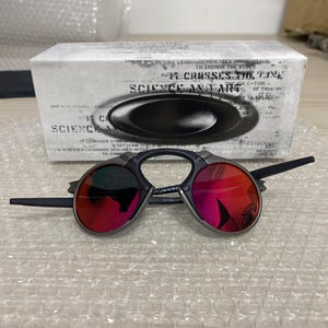 May include: Black sunglasses with red lenses are displayed in front of a white box. The sunglasses have a unique frame design. The box has the words "SCIENCE AND ART" printed on it.