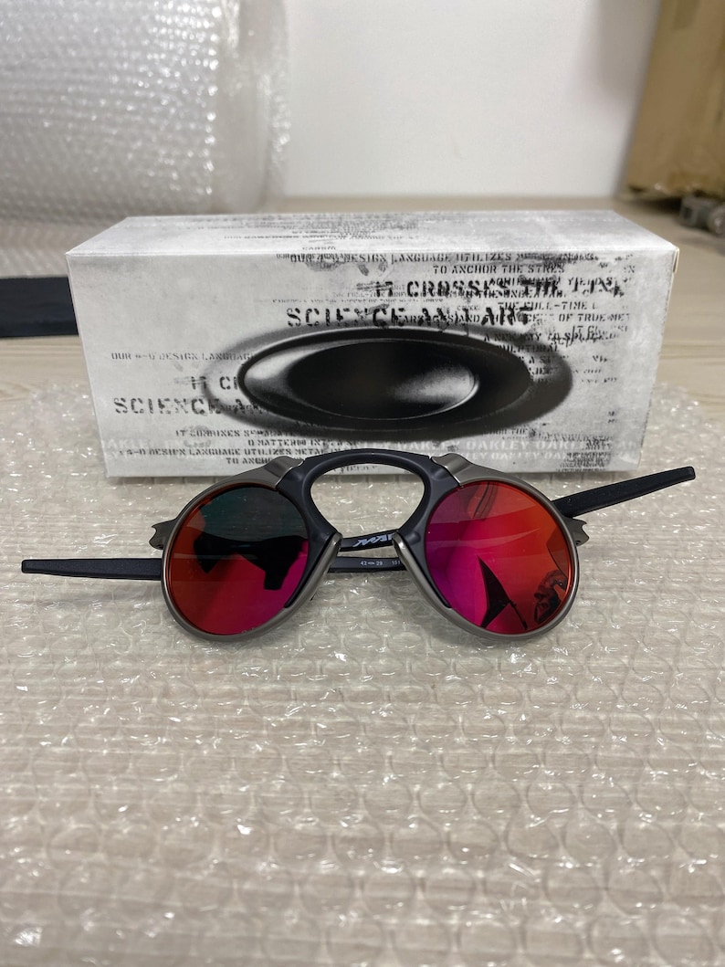 May include: Black sunglasses with red lenses are displayed in front of a white box. The sunglasses have a unique frame design. The box has the words "SCIENCE AND ART" printed on it.