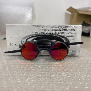 May include: Round sunglasses with red lenses and a black frame, featuring a silver bridge. The glasses are displayed on a white box with text. The overall design is modern and stylish.