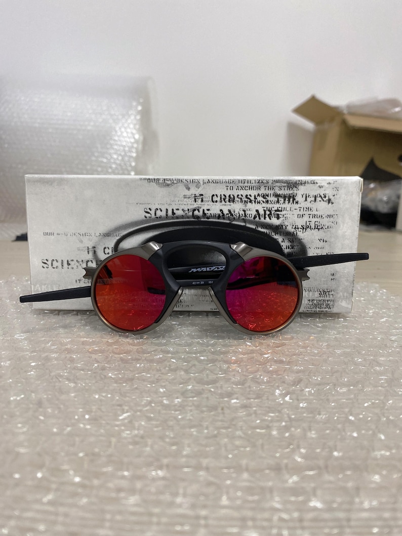 May include: Round sunglasses with red lenses and a black frame, featuring a silver bridge. The glasses are displayed on a white box with text. The overall design is modern and stylish.