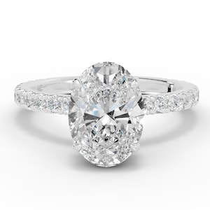 Peut inclure: Une bague de fiançailles en or blanc avec un grand diamant central de taille ovale. La bande est incrustée de petits diamants, créant un effet de halo autour de la pierre principale. La bague est sur fond blanc.